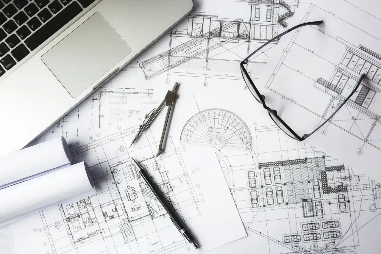 Na biurku leżą plany architektoniczne, laptop, cyrkiel, okulary i długopis. To miejsce pracy nad kosztorysem budowlanym.