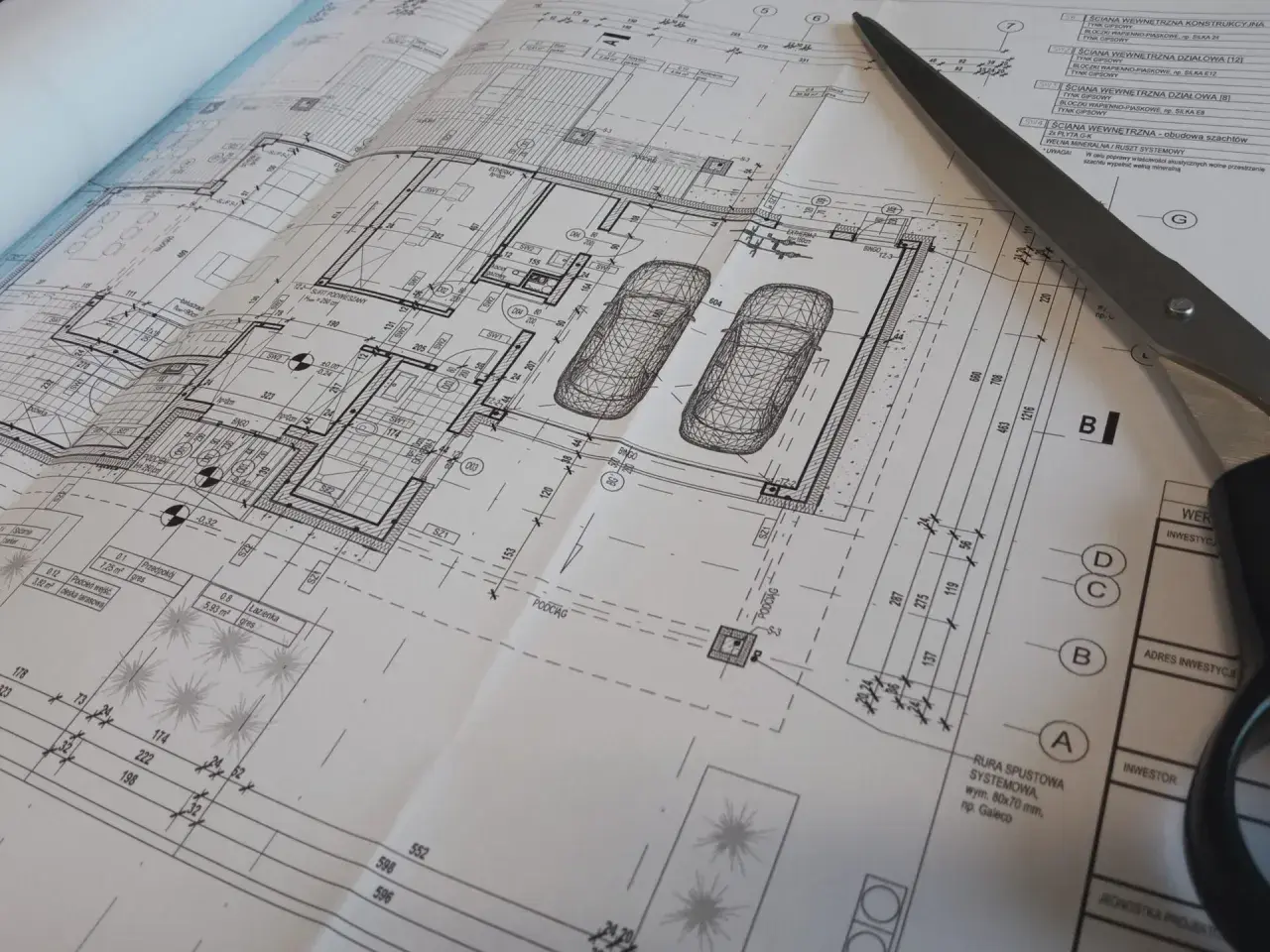 Szare nożyczki przecinają szczegółowy projekt budowlany domu z garażem.