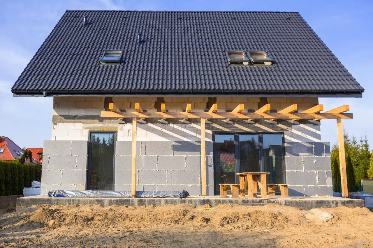 Budowa domu w toku, ściany z bloczków betonowych czekają na ocieplenie domu styropianem. Dach pokryty dachówką, widoczne okna dachowe i tarasowe.