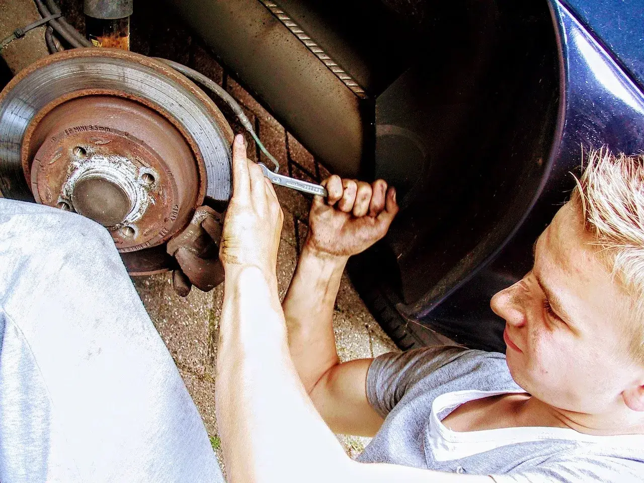 Młody mechanik z brudnymi rękami przykręca śrubę przy kole. Zastanawia się nad wymiana łożyska w kole cena.