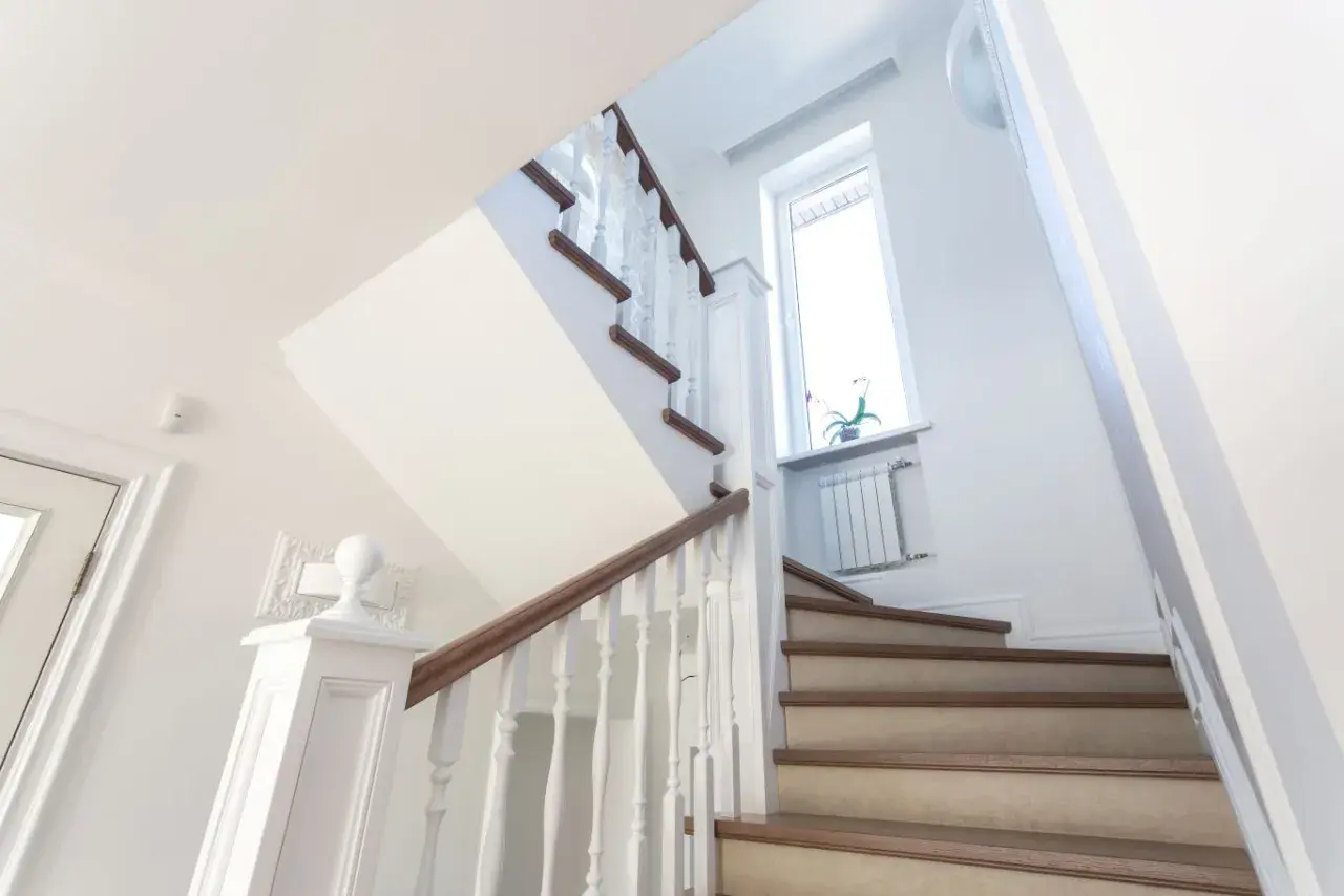 Jasne schody z drewnianymi stopniami i białymi balustradami. Inspiracja, jak tanio wykończyć schody betonowe, by wyglądały elegancko.