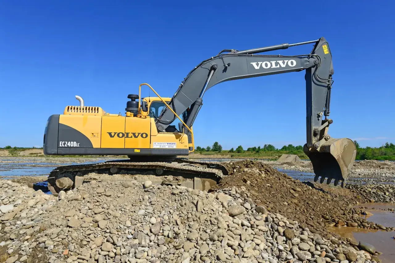 Żółta koparka Volvo EC240B LC pracuje na kamienistym brzegu rzeki. Zastanawiasz się, ile kosztuje godzina pracy koparki?