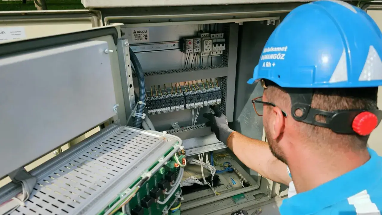 Elektryk w niebieskim kasku pracuje przy panelu sterowania. Potwierdza gotowość instalacji przyłączanej, dbając o każdy szczegół.
