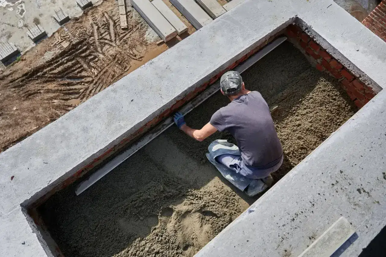 Budowlaniec wyrównuje świeży beton w wykopie. Widać elementy betonowe i ceglane.