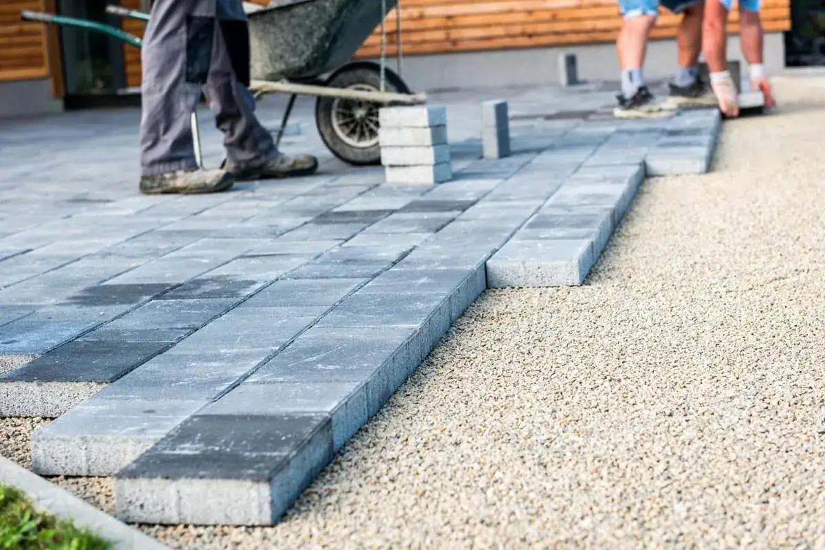 Układanie kostki brukowej na żwirze – świetny pomysł na tani podjazd. Dwie osoby pracują, jedna z taczką.