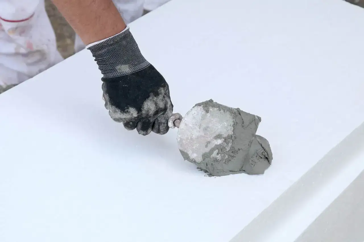 Ręka w rękawicy nakłada klej do styropianu na płytę. Określenie, ile kleju do styropianu na m2, jest kluczowe dla prawidłowego ocieplenia.