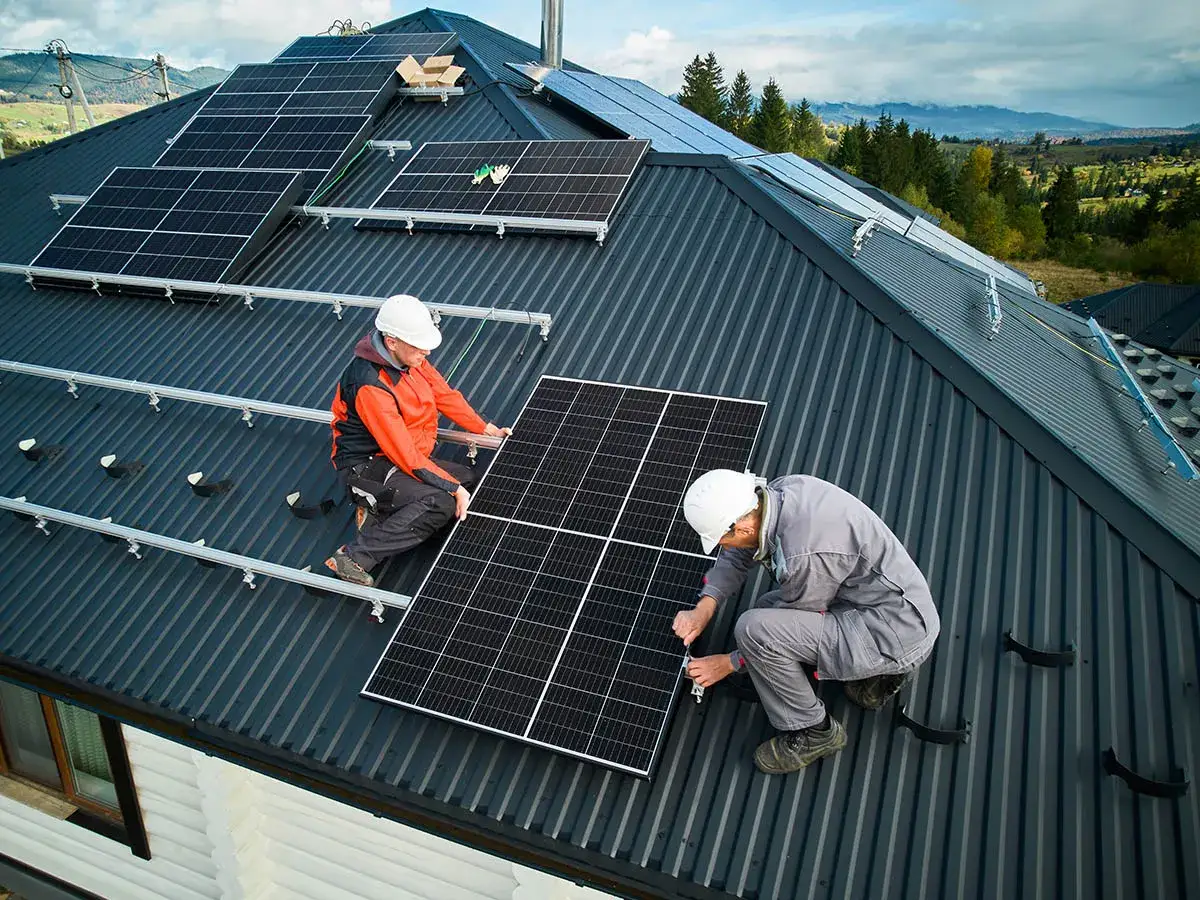 Dwóch pracowników w kaskach montuje panele fotowoltaiczne na dachu. Praca nad zieloną energią trwa.