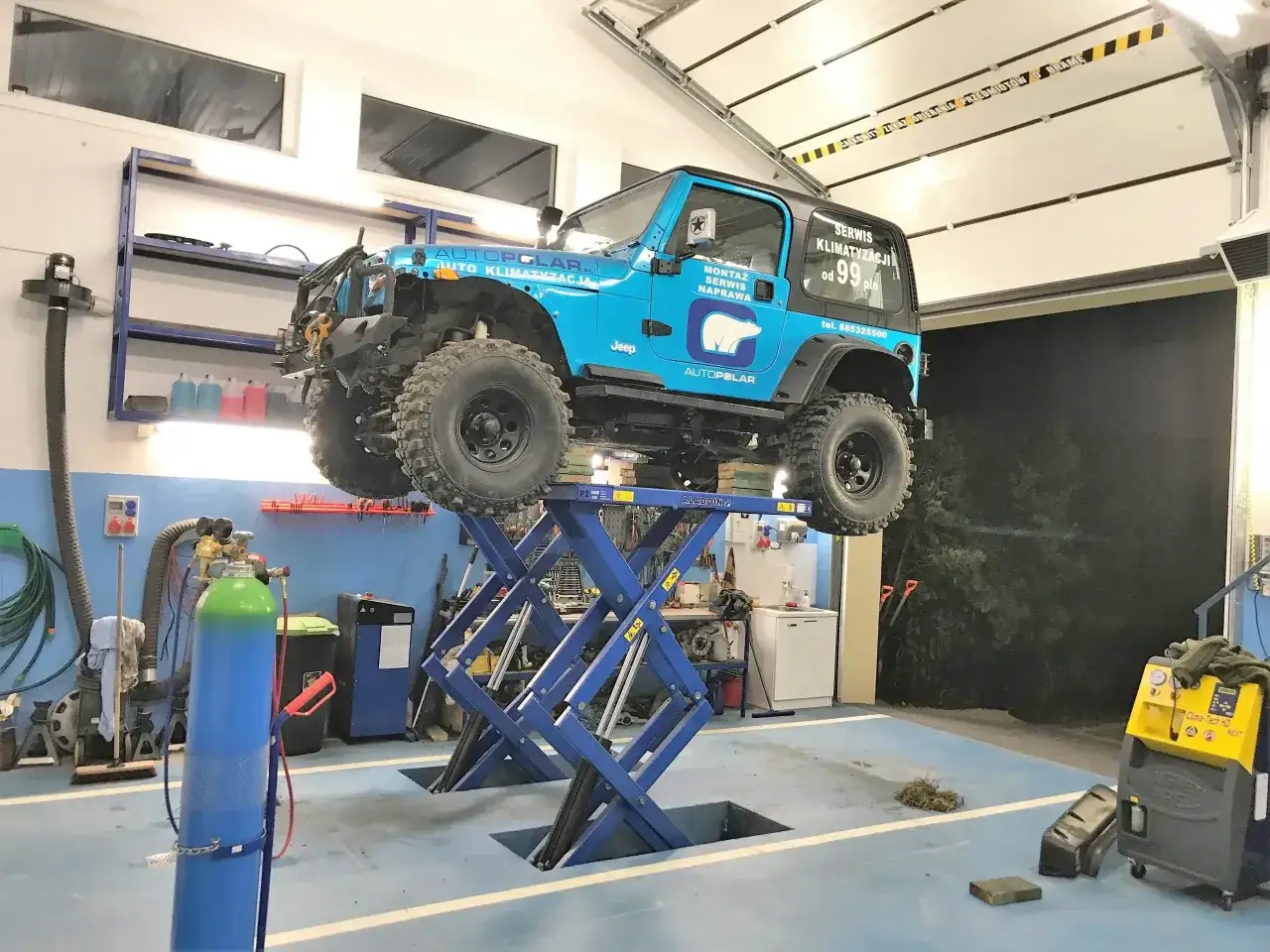 Niebieski Jeep na podnośniku w warsztacie. Na drzwiach informacja: