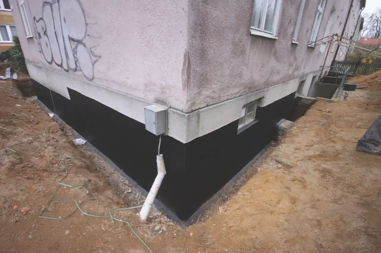 Prace przy izolacji fundamentów w starym domu. Czarna izolacja chroni ścianę przed wilgocią, a wykop odsłania ziemię wokół budynku.