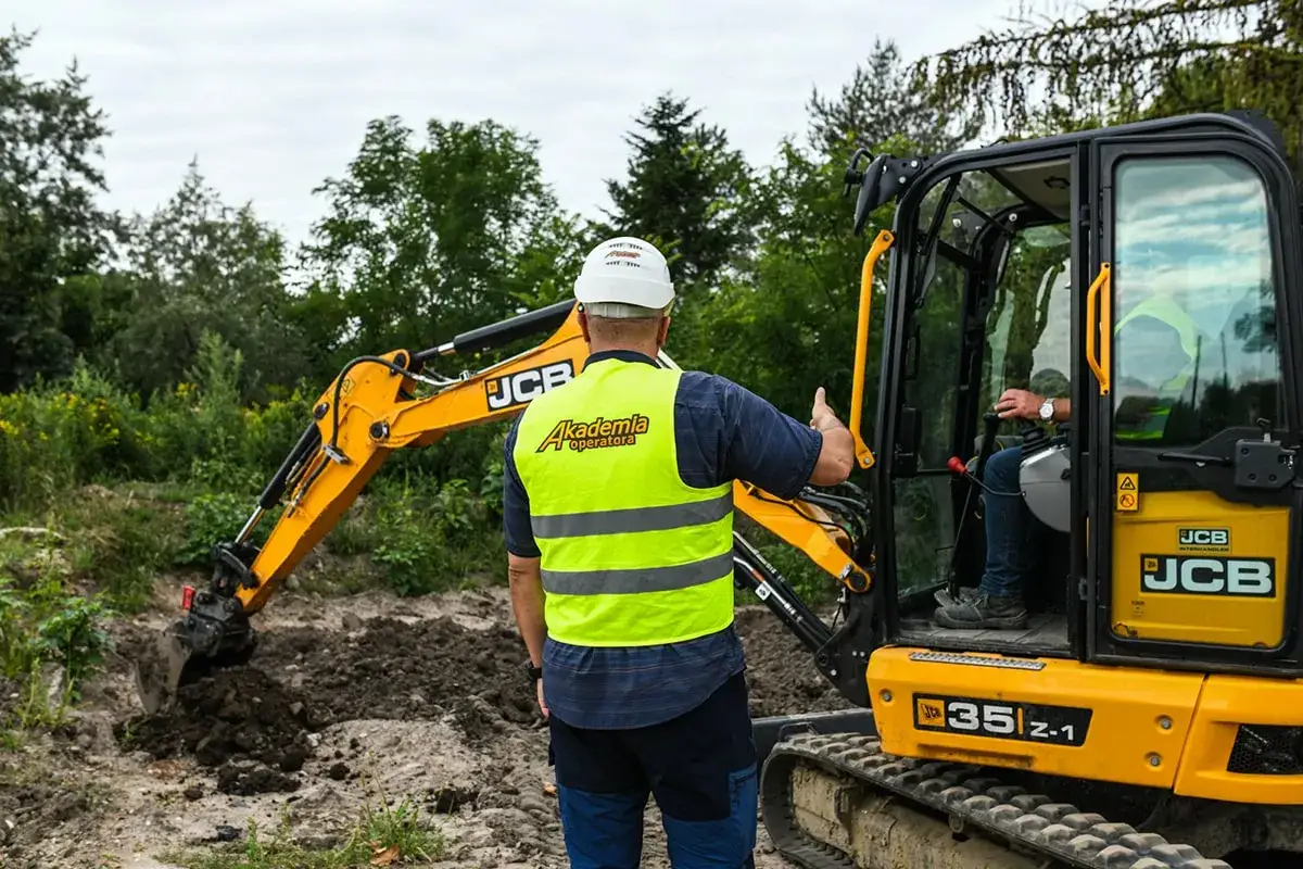 Operator w kamizelce odblaskowej instruuje, jak sprawdzić, ile kosztuje godzina pracy koparki. Maszyna JCB 35z-1 pracuje na budowie.